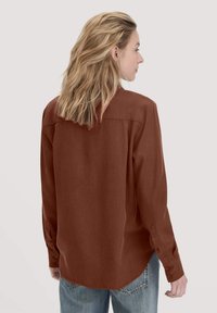 Camicia a maniche lunghe di colore ruggine, realizzata in tessuto liscio. Presenta polsini con bottoni e un design con orlo arrotondato, con yoke posteriore e dettagli di cucitura.