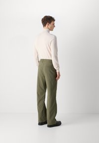 Gaišais beigu adījuma turtlenecks un olīvu zaļas plašās bikses ar smalku tekstūru. Modelis skatās prom, ģērbies melnās kurpēs.