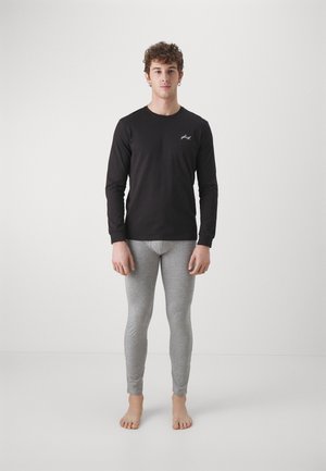 Cămașă neagră cu mânecă lungă, cu un logo alb pe piept, combinată cu colanți ajustați de culoare gri deschis, toate fabricate dintr-un material fin. Poziție pe picioarele goale.