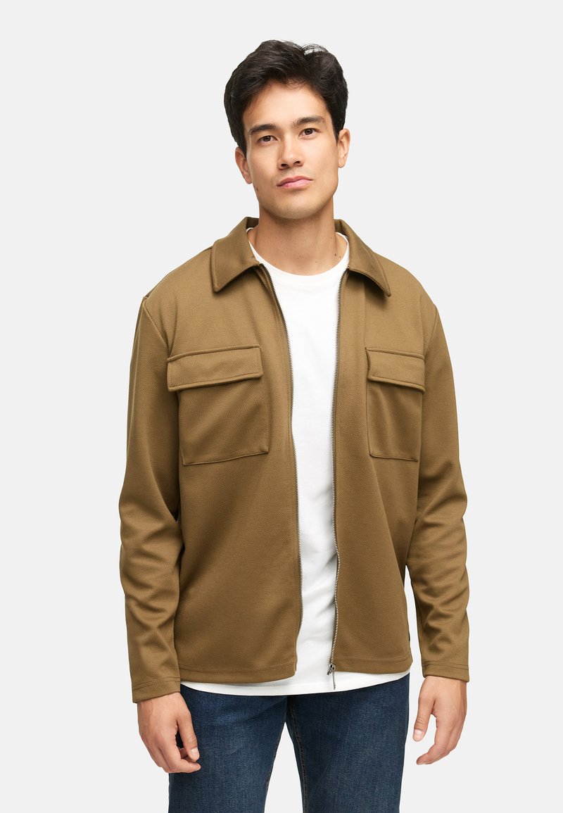 Brun lynlåsjakke med krave, to brystlommer og lange ærmer. Båret over en hvid skjorte, ser stoffet tekstureret og glat ud.