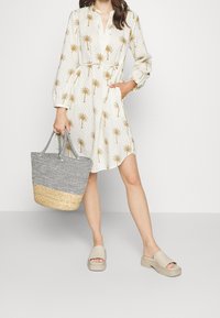 Vestido blanco con estampado de palmeras, cuello en V y mangas acampanadas; combinado con un bolso de mimbre en blanco y negro y sandalias de plataforma beige.