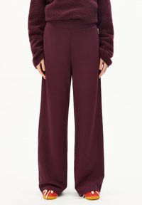 Hose mit weitem Bein aus tiefburgunderfarbenem Stoff, der eine glatte Textur und einen hohen Bund aufweist, kombiniert mit leuchtend orangen und gelben Schuhen.