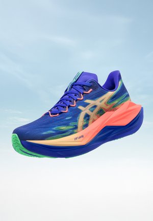 Blauer und grüner Laufschuh mit orangefarbener Zwischensohle, pinken Akzenten und Logo an der Seite, schwebend vor einem klaren Himmelshintergrund.