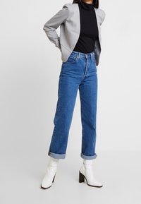 Harmaa rakennettu takki mustan korkean kauluksen paidan päällä, yhdistettynä vaaleansinisiin korkean vyötärön farkkuihin ja valkoisiin korollisiin nilkkasaappaisiin.