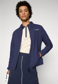 Navy zip-up jacket with a high collar, lightweight fabric, contrast stitching, and a logo on the left chest. Worn over a cream top.