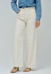 Jeans de pierna ancha en color crema con cintura alta, cinturilla y cierre de botón. Combinados con una camisa de mezclilla azul y tacones beige. Textura suave.