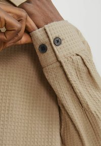 Kremo spalvos tekstūruotas marškinių rankogalis su vaflinio rašto ornamentu, dviejų juodų mygtukų ir papildomais raukšlėmis.