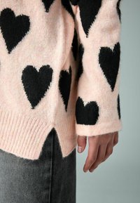 Rozā adīta džemperis ar lieliem melniem sirds rakstiem, ribotām piedurknēm un šķeltu apakšdaļu. Audums izskatās mīksts un teksturēts.