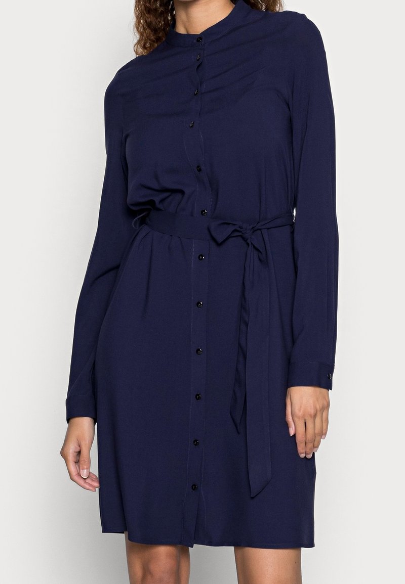 Femme portant une robe bleu marine à manches longues, boutonnée avec une ceinture à la taille, montrée des épaules jusqu'à mi-cuisse sur un fond uni.