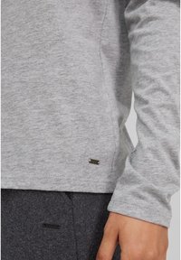 Graues Langarmshirt aus weichem Stoff mit leicht strukturiertem Finish. Besitzt ein kleines metallisches Logo-Tag in der Nähe des Saums.