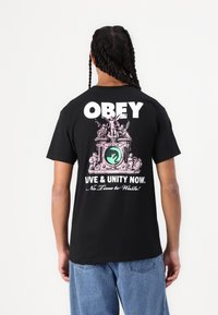 Svart bomull T-skjorte med stor hvit "OBEY" tekst, rosa grafisk design med et sentralt emblem, og teksten "KJÆRLIGHET & ENHET NÅ. Ingen tid å kaste bort!"
