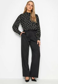 Svart blus med vita prickar, hög krage och lång ärm, kombinerad med skräddarsydda svarta byxor och svarta högklackade skor.