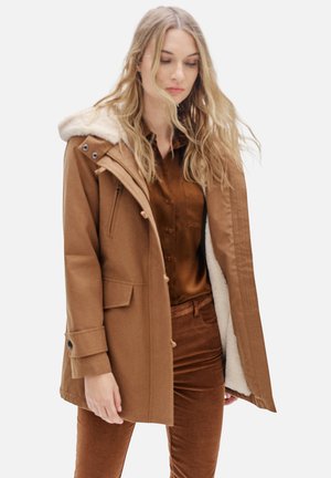 Veste parka marron avec capuche doublée de fausse fourrure, fermetures à boutons et zip, et poches latérales. Portée sur une chemise satinée marron à boutons et un pantalon.