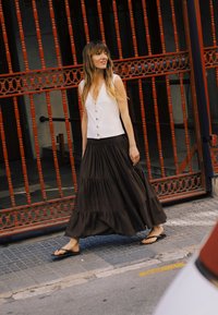 Femme aux longs cheveux portant un haut blanc sans manches, une jupe noire à volants et des tongs, marchant devant une porte métallique orange sur un trottoir.