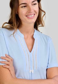 Blusa azul clara con mangas cortas, presenta un escote en V, detalles de encaje y un pequeño lazo blanco en la parte delantera. Tejido de textura suave.