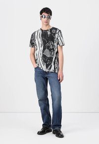 Svart-hvitt mønstrete t-skjorte med abstrakte design, kombinert med blå denimjeans og svarte sko. Modellen står med hendene i lommene.