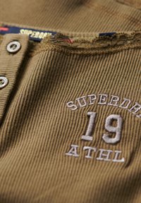 Shirt z ribbowanej tkaniny w oliwkowym kolorze, z kołnierzykiem o surowych krawędziach, z srebrnymi guzikami i białym haftowanym napisem: „SUPERDRY 19 ATHL.”