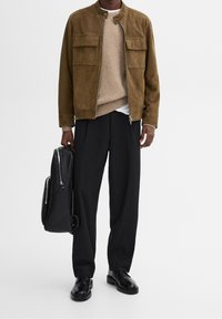 Giacca in suede marrone con due tasche sul petto, maglione in maglia beige, pantaloni neri e zaino in pelle nera. Le scarpe nere completano l'outfit.