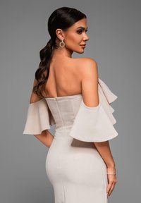 Světlé béžové off-shoulder šaty s rozšířenými rukávy, přiléhavým korzetem a hladkou texturou. Stříbrné kruhové náušnice zvyšují eleganci.