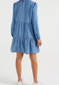 Denimklänning i ljusblått med långa puffärmar, lager-på-lager volanger och avslappnad passform. Inkluderar rund halsringning och elastiska manschetter.