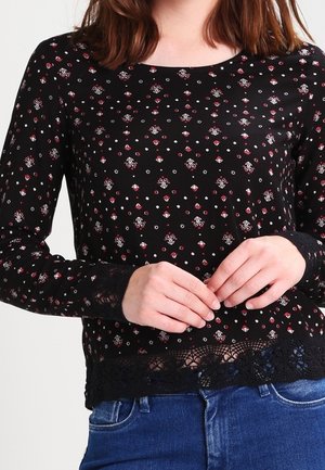 Kobieta ubrana w czarną bluzkę z długim rękawem ozdobioną małymi czerwono-białymi kwiatowymi wzorami, z koronkowymi wykończeniami na mankietach i dole, w połączeniu z niebieskimi dżinsami.