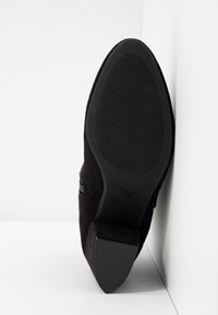 Bottine en suède noir avec un bout effilé et une semelle en caoutchouc texturée. Elle comporte un petit talon et une fermeture éclair latérale pour un enfilage facile.