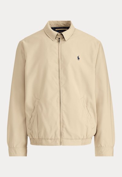 Casaco leve bege com gola de colarinho, fecho de correr frontal, punhos elásticos e dois bolsos laterais. Apresenta um pequeno logótipo escuro no peito.