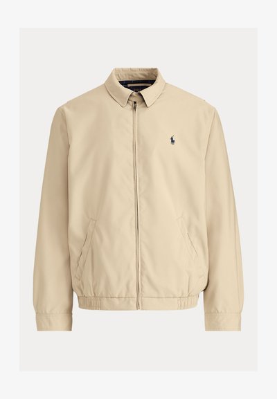 Casaco leve bege com gola de colarinho, fecho de correr frontal, punhos elásticos e dois bolsos laterais. Apresenta um pequeno logótipo escuro no peito.