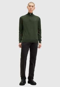 Žalias megztinis su užtrauktuku prie kaklo, aptimaus silueto, pagamintas iš lygios medžiagos. Dėvimas su juodais džinsais ir tvirtomis juodomis batais.