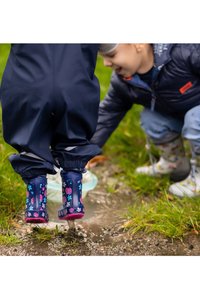 Blauwe rubberen laarzen met roze lieveheersbeestje- en bloemenpatronen; gecombineerd met donkerblauwe waterdichte overall, geplaatst in een grasgebied bij water.