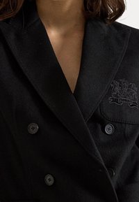 Närbild av en person som bär en svart dubbelknäppt kappa med broderad Ralph Lauren-logotyp på bröstfickan.