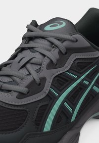 Σκούρο γκρι και μαύρο αθλητικό παπούτσι με τόνους τυρκουάζ, λεπτομερείς ραφές και σφιχτά δεμένα γκρι κορδόνια σε λευκό φόντο.