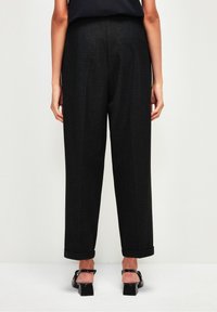 Mustat muotoon leikattut housut suoran lahkeiden kanssa, joissa on käärityt lahkeet ja hienovarainen tekstuuri. Yhdistetty tumman päällystakin ja hihnallisten korkokenkien kanssa.