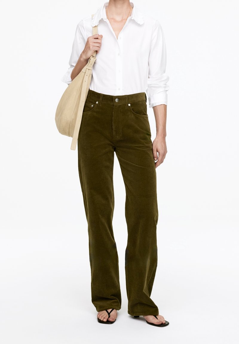 Femme portant un pantalon en velours côtelé vert olive, une chemise blanche boutonnée, un sac beige à l'épaule et des sandales noires à brides, debout devant un fond blanc.