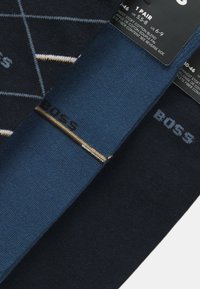 Due coppie di calzini: una blu navale con un motivo geometrico e l'altra blu scuro uniforme, entrambe con il marchio "BOSS" in colori a contrasto.