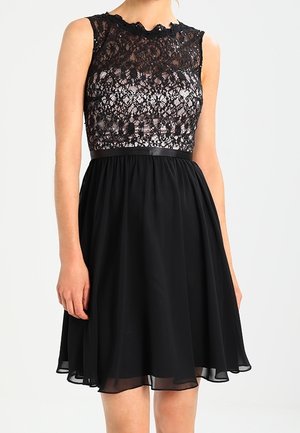 Robe noire avec un corsage en dentelle à encolure festonnée, taille cintrée et jupe fluide en chiffon. Détail simple d'une ceinture noire.