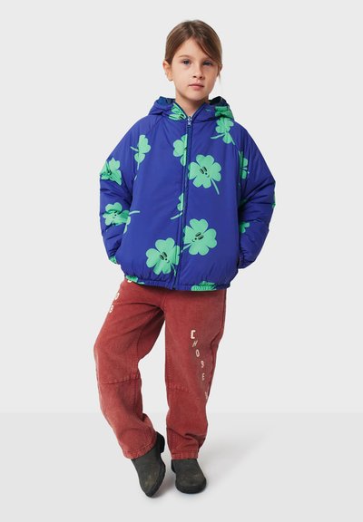 Veste matelassée bleue avec un motif floral vert et une capuche. Associée à un pantalon rouge ample avec un texte blanc. Bottines noires à la cheville.