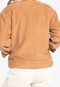 Veste en velours côtelé couleur fauve avec texture côtelée et col doublé de fourrure ; manches longues et ourlet court, associée à un pantalon blanc.