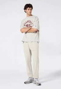 Lichtgrijs t-shirt met bordeauxrode tekst, gecombineerd met lichtgrijze joggingbroek. Sneakers zijn grijs met witte accenten. Eenvoudig ontwerp met een relaxte pasvorm.