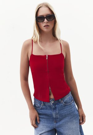 Woman wearing black sunglasses, red spaghetti strap top with front zipper, and blue high-waisted jeans against a white background.