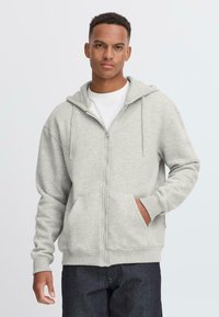 Grey zip-up hoodie made of soft fabric, featuring a drawstring hood, kangaroo pockets, and ribbed cuffs. Worn over a white shirt.