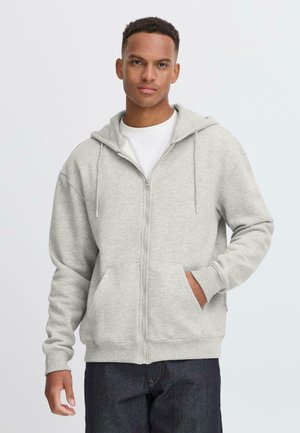 Grå zip-up hoodie lavet af blødt stof, med en hætte med snoretræk, kængurulommer og ribbede manchetter. Båret over en hvid skjorte.
