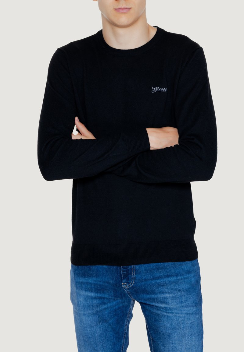Maglione nero a maglia con maniche lunghe, collo rotondo e discreto logo sul petto. Abbinato a jeans in denim blu chiaro.