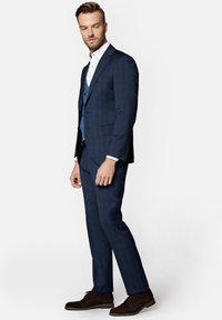 Granatowy, w kratkę, dopasowany garnitur, zestawiony z białą koszulą i niebieską kamizelką. Brązowe eleganckie buty dopełniają całość.