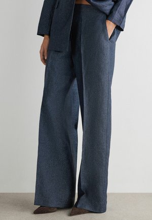 Kvinde iført navyblå bukser med vide ben og lommer, en matchende blazer og brune vævede sko med spids tå, stående mod en ensfarvet baggrund.