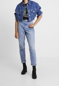 Denimjakke i mellemblå, cropped-design med to brystlommer. Båret over en sort grafik t-shirt, parret med højtaljede lyseblå jeans og sorte støvler.