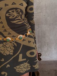 Zelený a hnedý vzorovaný sveter s priliehavým strihom, doplnený dekoratívnym koženým opaskom s obručovými zelenými akcentmi.