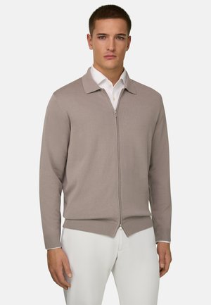 Jeune homme portant un cardigan beige à fermeture éclair par-dessus une chemise blanche à col et un pantalon blanc, debout devant un fond uni.