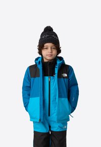 Modrá a čierna termo bunda s kapucňou, so zipsom vpredu, doplnená o zodpovedajúce modré nohavice. Dieťa má na hlave čiernu pletenú čiapku s brmbolcom.