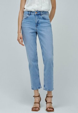 Person trägt hellblaue High-Waist-Jeans mit geradem Bein, ein weißes Oberteil und braune Riemchen-Sandaletten mit Absatz, steht vor einem einfarbigen Hintergrund.
